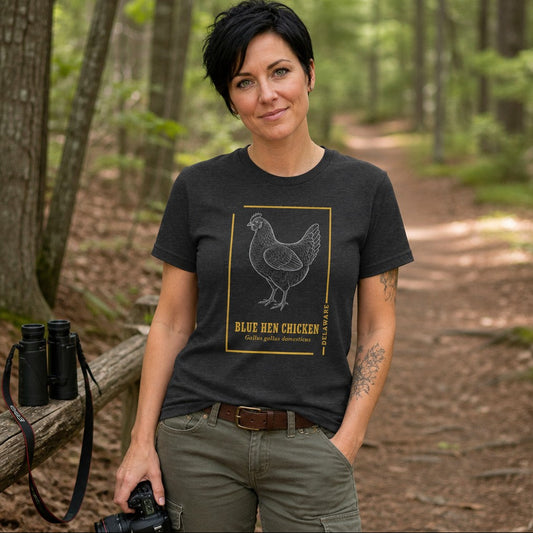 Woman wearing a dark grey heather Bella Canvas 3001 t-shirt featuring a vintage-style line art illustration of the Blue Hen Chicken in a gold border with botanical accents, standing on a wooded trail, ideal for birdwatchers and wildlife enthusiasts, cottagecore heritage design, by Crossroads Dandelion