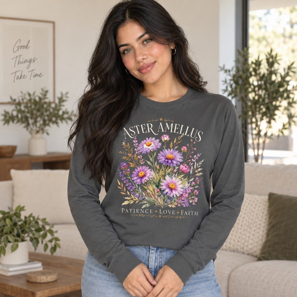 Woman wearing an asphalt Bella Canvas® 3501 long sleeve featuring a vintage botanical design of hand-painted aster and amellus flowers in soft lavender, pink, and violet hues with golden wheat accents, cream serif 'ASTER AMELLUS' and 'PATIENCE LOVE FAITH', romantic cottagecore floral apparel for nature lovers, indoor living room, by Crossroads Dandelion