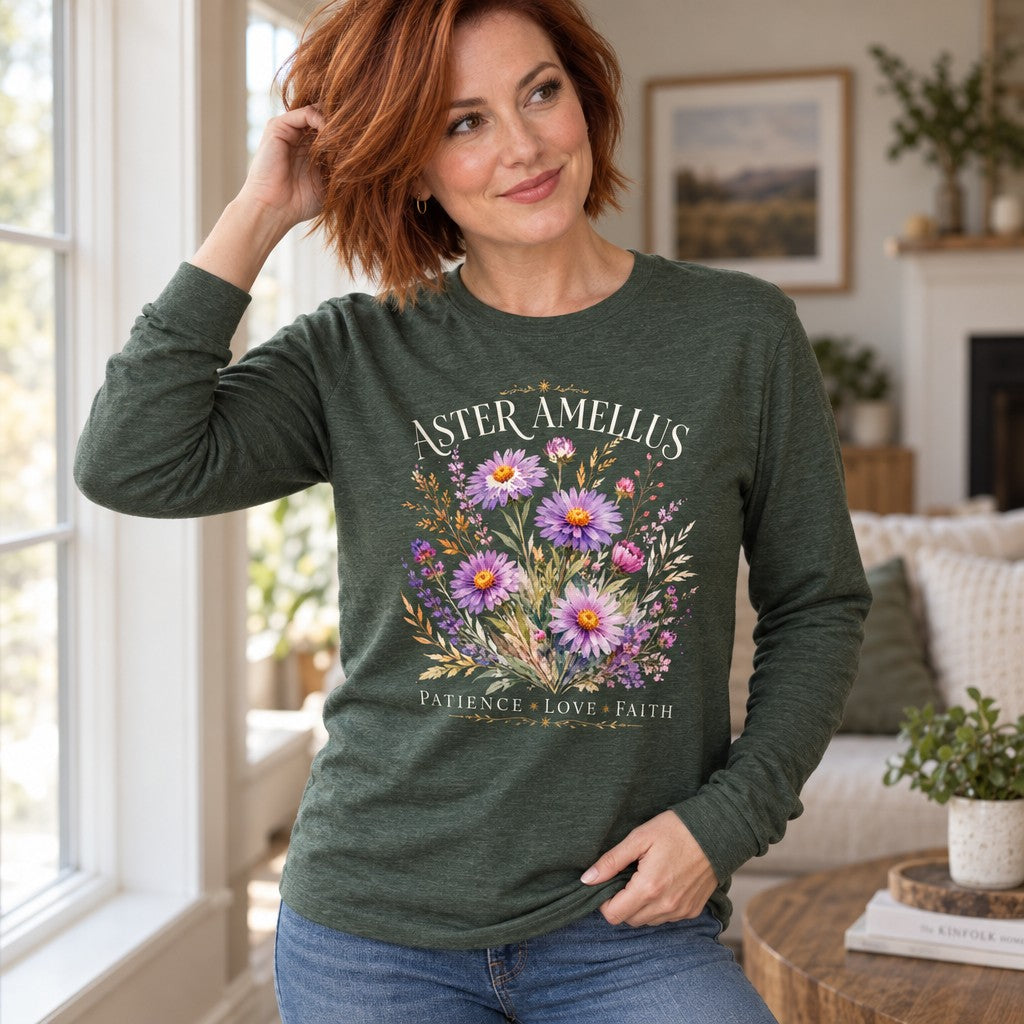 Woman wearing a heather forest Bella Canvas® 3501 long sleeve shirt with a vintage botanical bouquet of hand-painted aster and amellus flowers in soft lavender and pink hues, golden wheat accents, cream serif text ASTER AMELLUS and inspirational words, cozy cottage interior, romantic floral apparel for nature lovers, by Crossroads Dandelion
