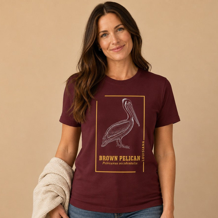 Woman in maroon Bella Canvas 3001 t-shirt featuring a vintage engraved brown pelican illustration with golden ochre framing, classic heritage birdwatcher and naturalist gift, by Crossroads Dandelion