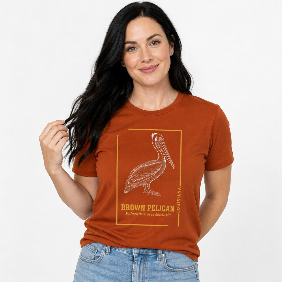 Woman in autumn orange Bella Canvas 3001 t-shirt featuring a vintage engraving style brown pelican illustration with golden ochre framing, classic contemplative mood for birdwatchers and heritage enthusiasts, by Crossroads Dandelion