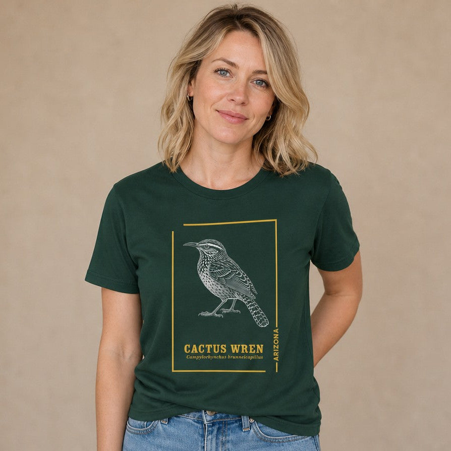 Woman wearing a forest green Bella Canvas 3001 t-shirt with a detailed vintage line art illustration of a Cactus Wren, Arizona state bird, outlined with golden accents and botanical-inspired text, perfect for bird lovers, southwest enthusiasts, and nature fans, by Crossroads Dandelion
