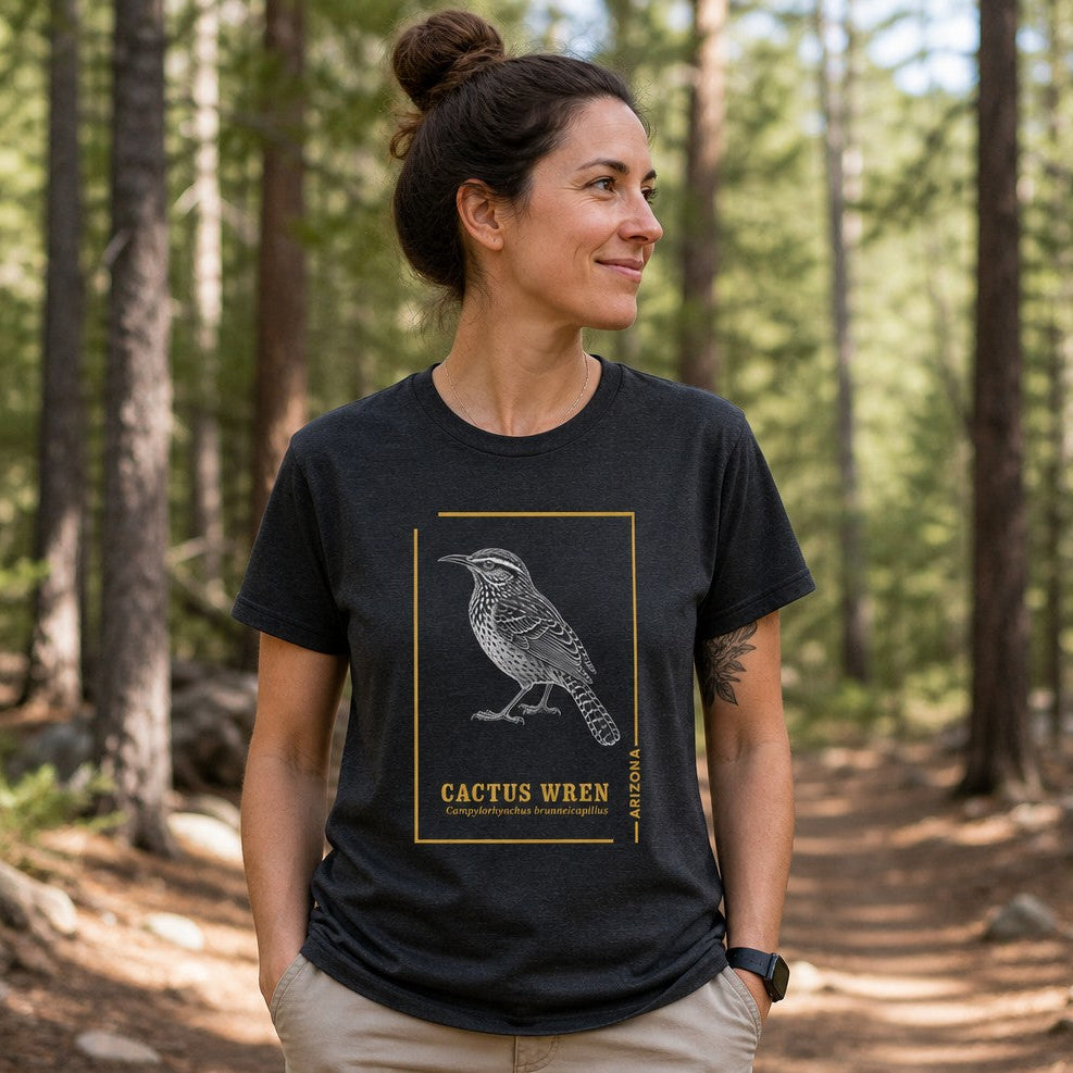 Woman wearing a dark grey heather Bella Canvas t-shirt featuring a vintage line art illustration of the Cactus Wren, Arizona state bird, with golden frame and botanical-inspired type, standing on a forest trail, perfect for ornithology and Southwest nature lovers, by Crossroads Dandelion
