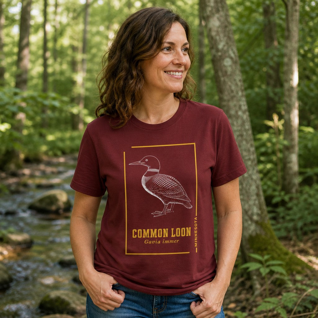 Woman in cardinal red Bella Canvas 3001 t-shirt with a vintage-style black and white etching of a common loon, detailed feather illustration framed in gold, cottagecore and wildlife lover design, standing in a forest by a stream, perfect for birdwatchers and nature enthusiasts, by Crossroads Dandelion