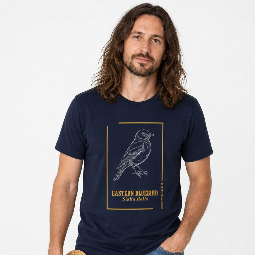 Man wearing navy Bella Canvas 3001 t-shirt featuring a detailed white line art Eastern Bluebird illustration with golden vintage botanical framing and scientific name, for birdwatchers and nature lovers, by Crossroads Dandelion