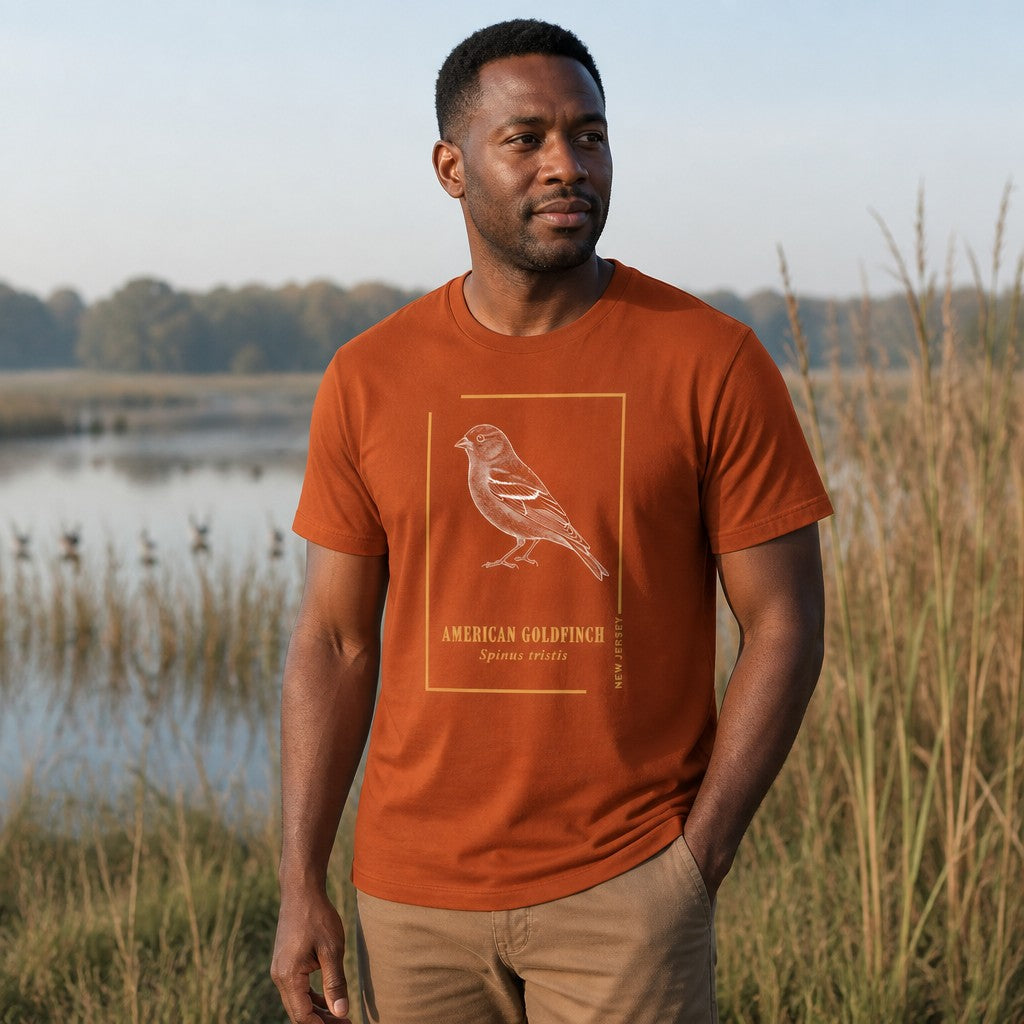Man wearing an autumn orange Bella Canvas 3001 t-shirt featuring a vintage etching style illustration of the American Goldfinch within a golden frame, classic field guide aesthetic, perfect for birdwatchers and nature lovers, outdoors by the water, men's apparel, by Crossroads Dandelion