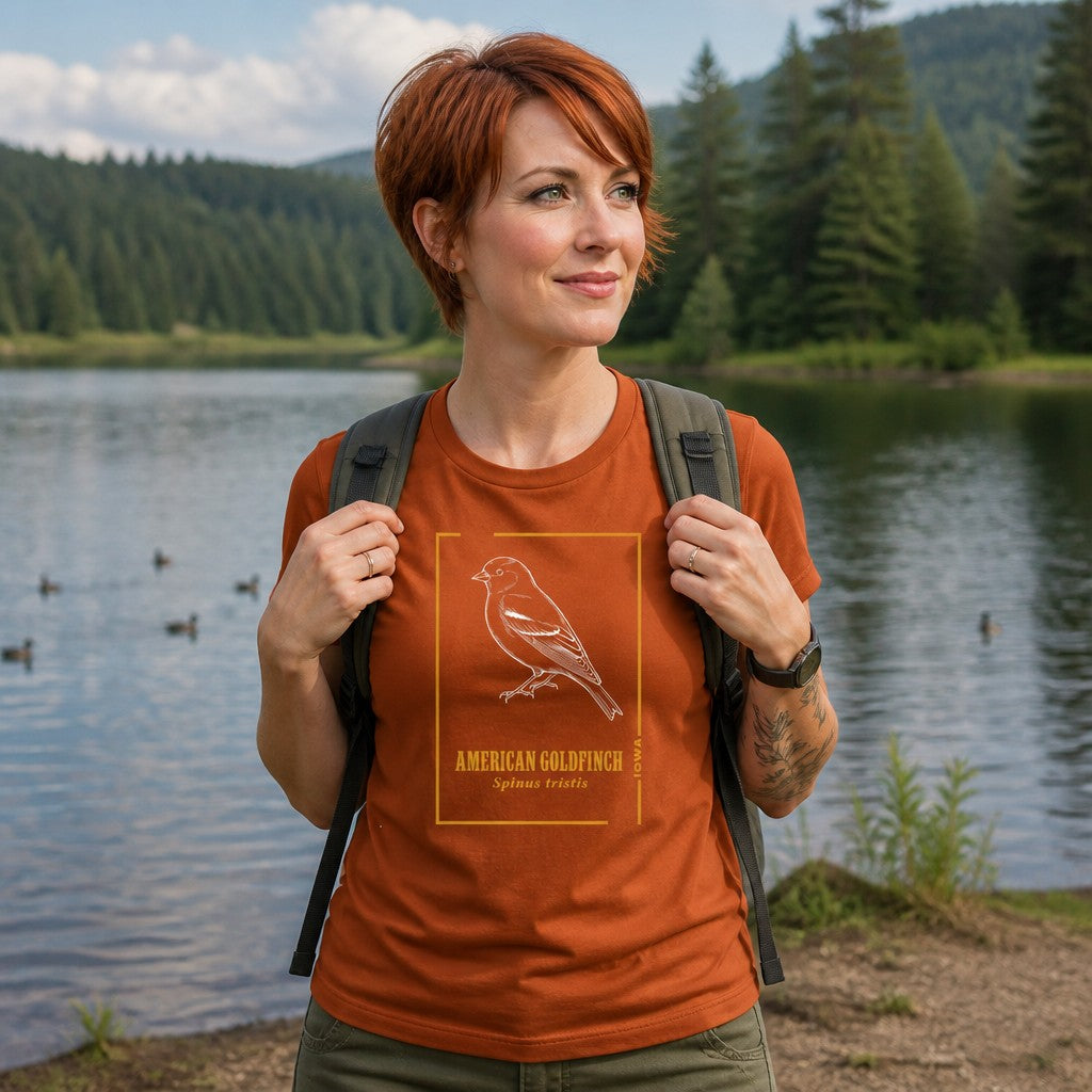 Woman wearing an autumn orange Bella Canvas 3001 t-shirt with a vintage-style line art American Goldfinch design, hand-drawn and framed by botanical accents, walking by a serene lake with pine trees, ideal for bird and nature lovers, by Crossroads Dandelion