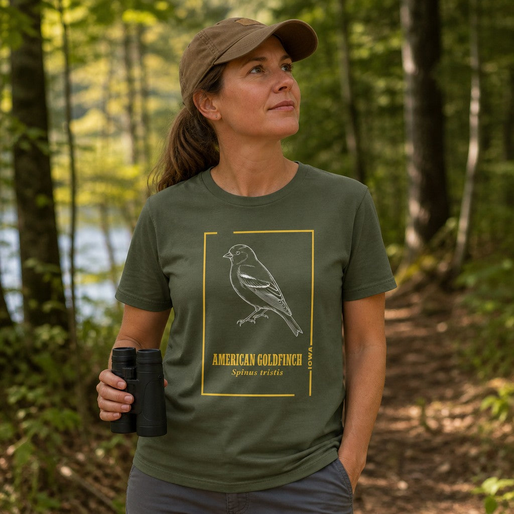 Woman wearing a military green Bella Canvas 3001 t-shirt with a vintage hand-drawn line art illustration of an American Goldfinch framed by golden botanical accents, perfect for birding enthusiasts and nature lovers, hiking in a wooded outdoor setting, by Crossroads Dandelion