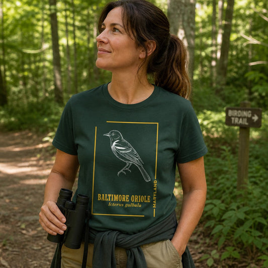 Woman wearing a forest green Bella Canvas 3001 t-shirt with a vintage-inspired white line art illustration of a Baltimore Oriole, Maryland state bird, framed in golden yellow, hand-drawn field guide style, hiking on a birding trail, ideal for birdwatchers and nature enthusiasts, by Crossroads Dandelion