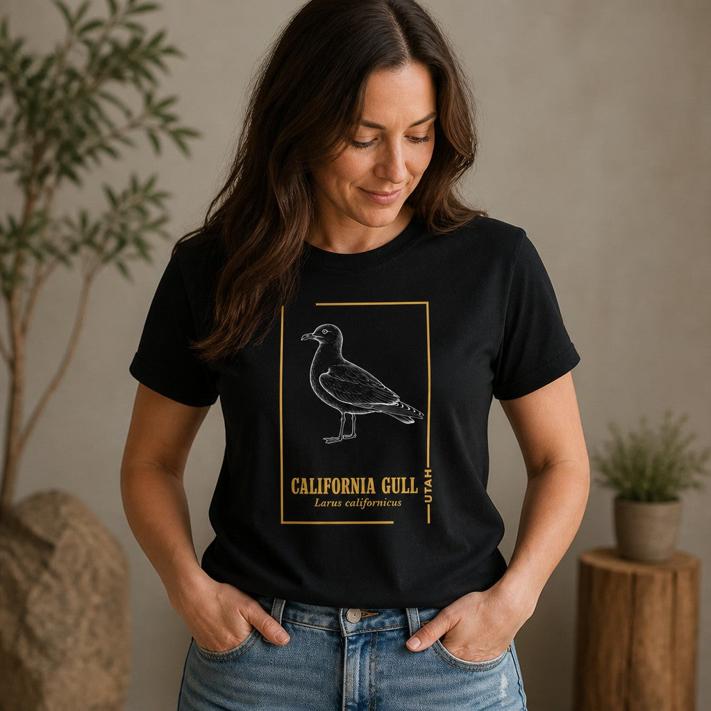 Woman in black Bella Canvas 3001 t-shirt featuring a vintage-style line art illustration of a California Gull with golden frame and botanical accents, designed for birdwatchers and nature lovers, photographed indoors, by Crossroads Dandelion