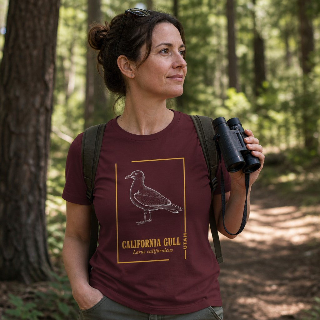 Woman wearing a maroon Bella Canvas 3001 t-shirt with a vintage line art California Gull design in chalk-white with gold script, hiking in a forest, perfect for birdwatchers, naturalists, and wildlife art lovers, by Crossroads Dandelion