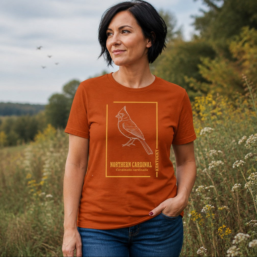 Woman in autumn orange Bella Canvas 3001 t-shirt featuring a vintage-inspired white line illustration of a Northern Cardinal with botanical typography, standing in a wildflower field, classic wildlife and birdwatching lovers, by Crossroads Dandelion