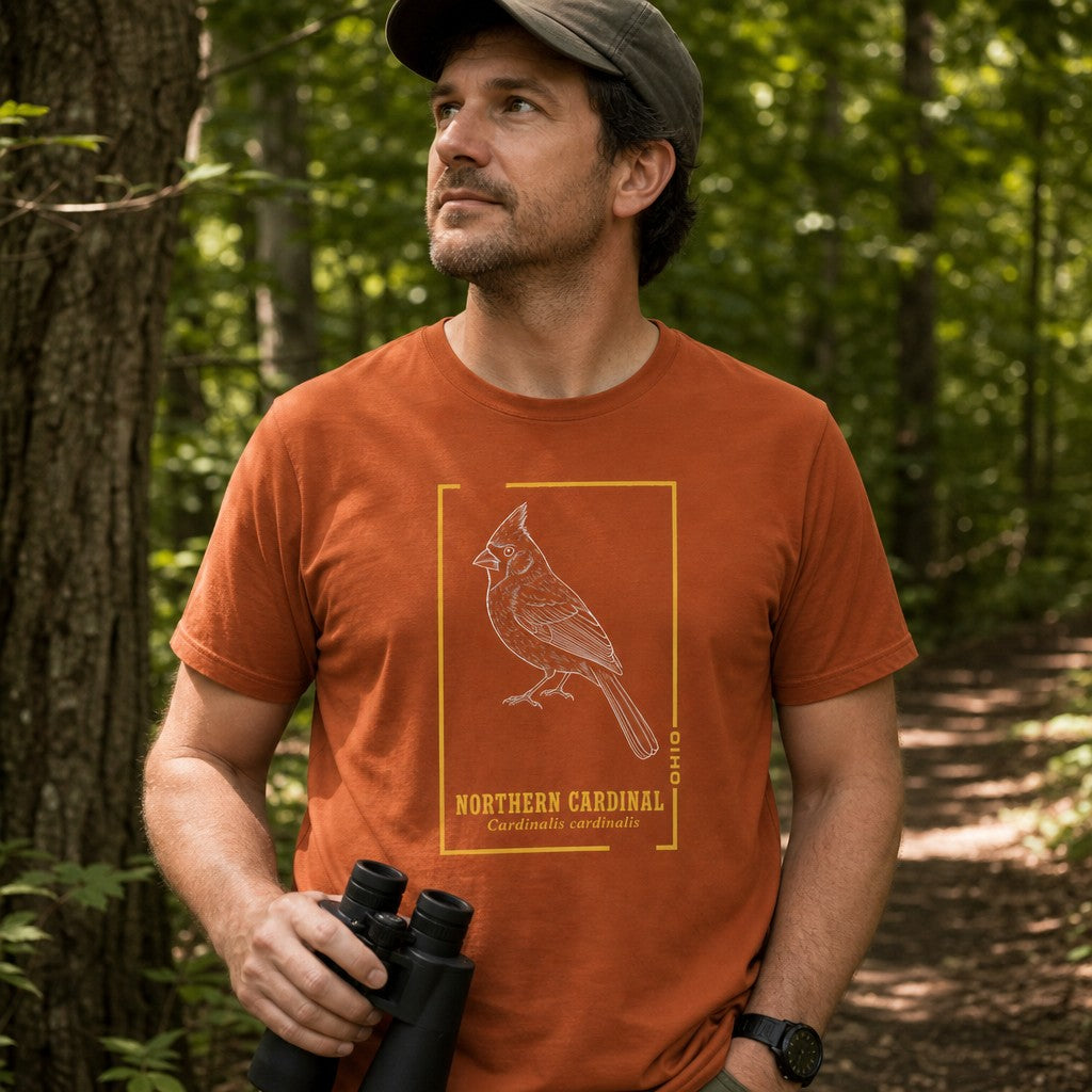 Man in an autumn orange Bella Canvas 3001 t-shirt featuring a vintage-inspired hand-drawn white line art Northern Cardinal within a gold botanical frame and field guide-style lettering, hiking in a wooded forest, perfect for birdwatchers and wildlife enthusiasts, by Crossroads Dandelion