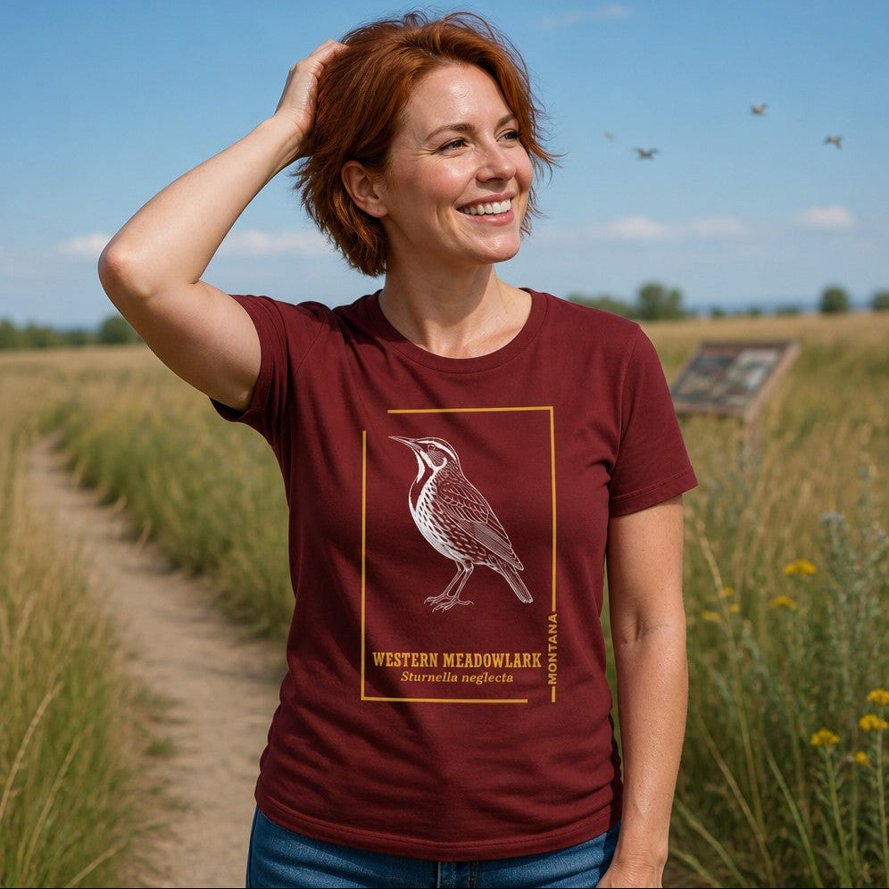 Woman wearing a cardinal Bella Canvas 3001 t-shirt featuring a hand-drawn vintage etching of a Western Meadowlark in profile, framed with golden lines and elegant typography, perfect for birdwatchers and heritage nature art enthusiasts, photographed in a grassy outdoor setting, by Crossroads Dandelion