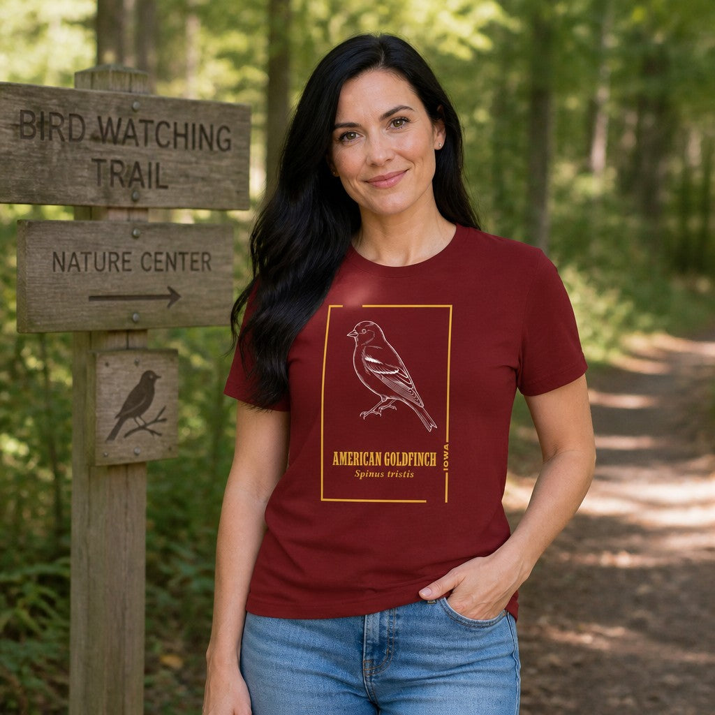 Woman wearing cardinal red Bella Canvas 3001 t-shirt with vintage line art illustration of an American Goldfinch and golden botanical accents, standing at a bird watching trail in the woods, ideal for bird lovers and nature enthusiasts, by Crossroads Dandelion