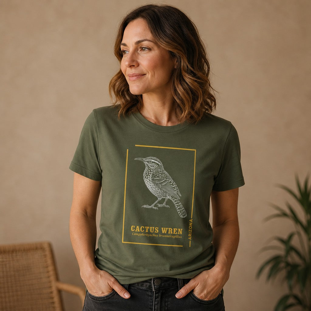 Woman wearing military green Bella Canvas 3001 t-shirt with vintage-style line art illustration of a Cactus Wren, Arizona state bird, surrounded by golden botanical accents, indoor casual scene, perfect for nature enthusiasts, bird watchers, and lovers of Southwest heritage, by Crossroads Dandelion