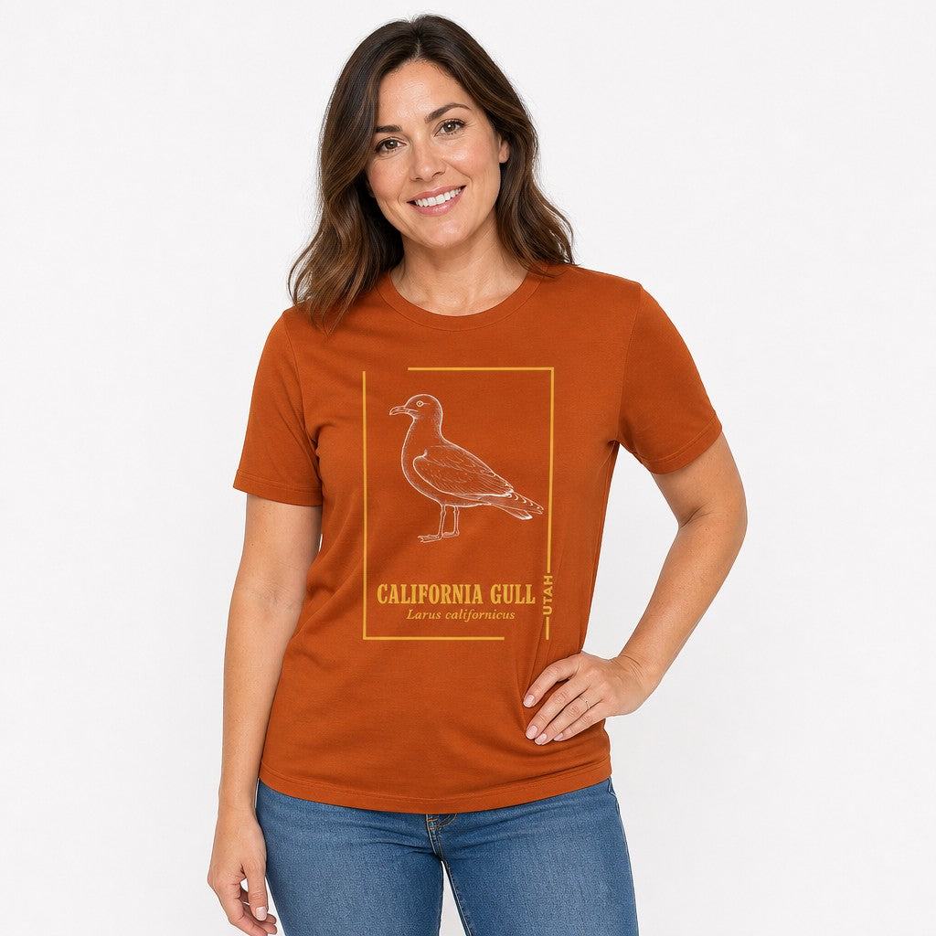 Woman wearing an autumn orange Bella Canvas 3001 t-shirt featuring a vintage-style line art illustration of the California Gull with golden botanical accents, ideal for birdwatchers and lovers of classic wildlife art, by Crossroads Dandelion
