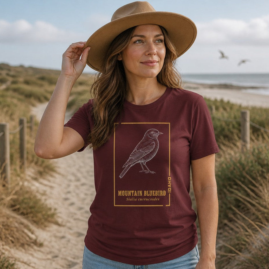 Woman wearing maroon Bella Canvas 3001 t-shirt with vintage line art Mountain Bluebird design, botanical golden accents and scientific name, birdwatcher and nature lover gift, coastal dunes stroll, by Crossroads Dandelion