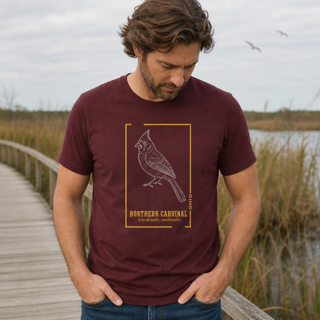 Man wearing a maroon Bella Canvas 3001 t-shirt featuring a vintage-inspired white line art illustration of a Northern Cardinal in a golden botanical frame, bold serif and italic text, standing on a wooden boardwalk in a nature reserve, perfect for birdwatchers and vintage wildlife lovers, by Crossroads Dandelion