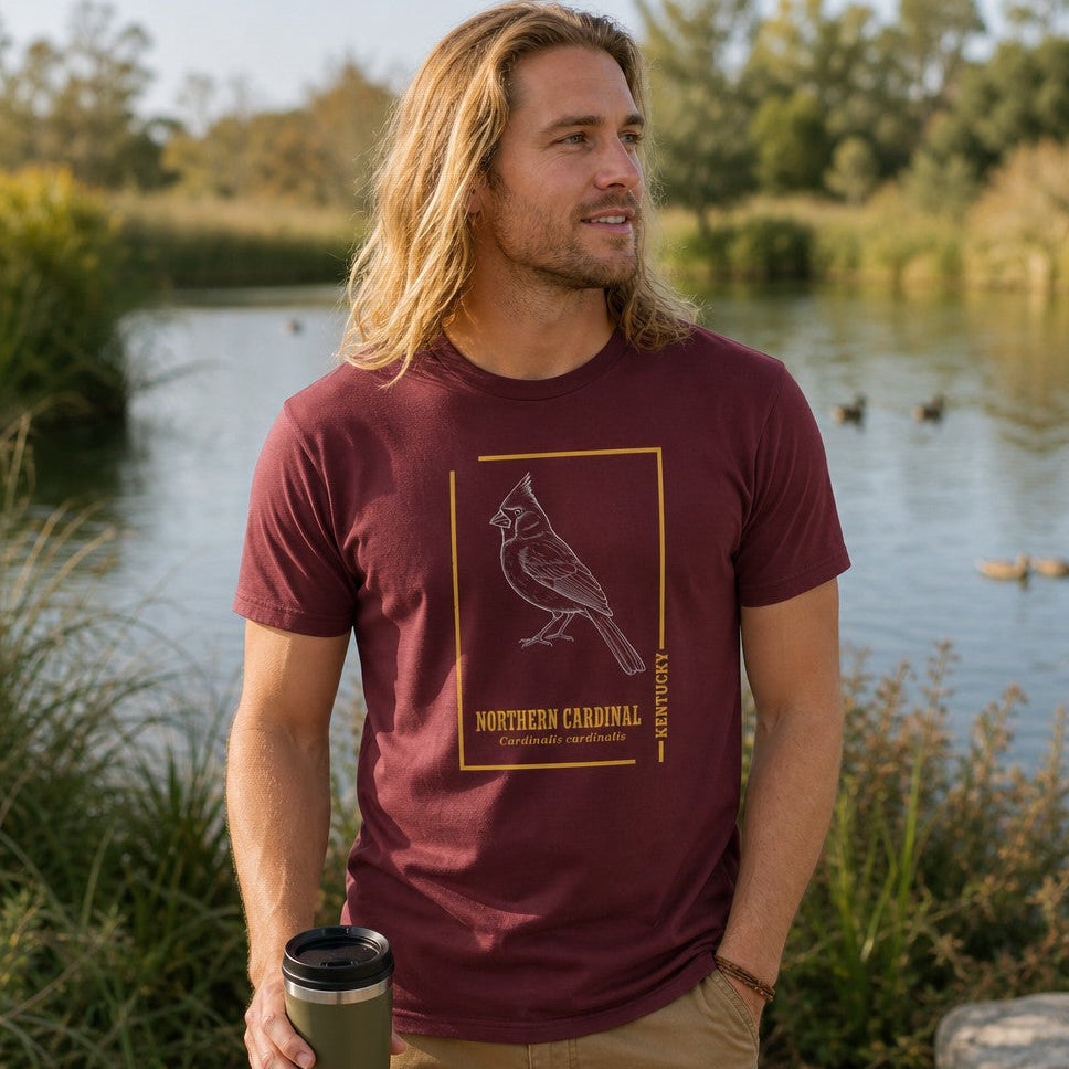 Man in maroon Bella Canvas 3001 t-shirt featuring a vintage-inspired white line art illustration of a Northern Cardinal with gold border and botanical typography, standing by a lakeside, perfect for birdwatchers and nature lovers, by Crossroads Dandelion