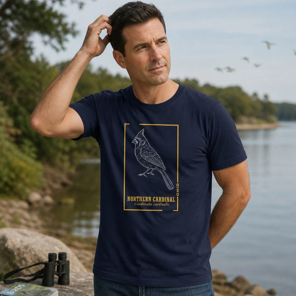 Man in navy Bella Canvas 3001 t-shirt featuring a vintage-inspired white line art illustration of a Northern Cardinal framed in gold, styled like a field guide, outdoors by a scenic lake, perfect for birdwatchers and nature lovers, by Crossroads Dandelion