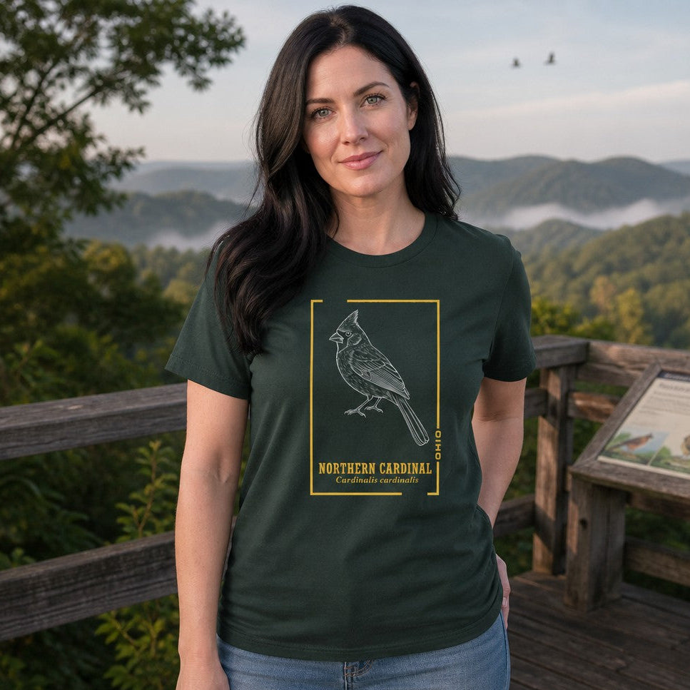 Woman wearing a forest green Bella Canvas tee featuring a vintage hand-drawn white line art illustration of a Northern Cardinal inside a golden botanical frame, with bold serif and italicized Latin text, standing at a scenic overlook with mountain views, perfect for female birdwatchers and vintage wildlife enthusiasts, by Crossroads Dandelion
