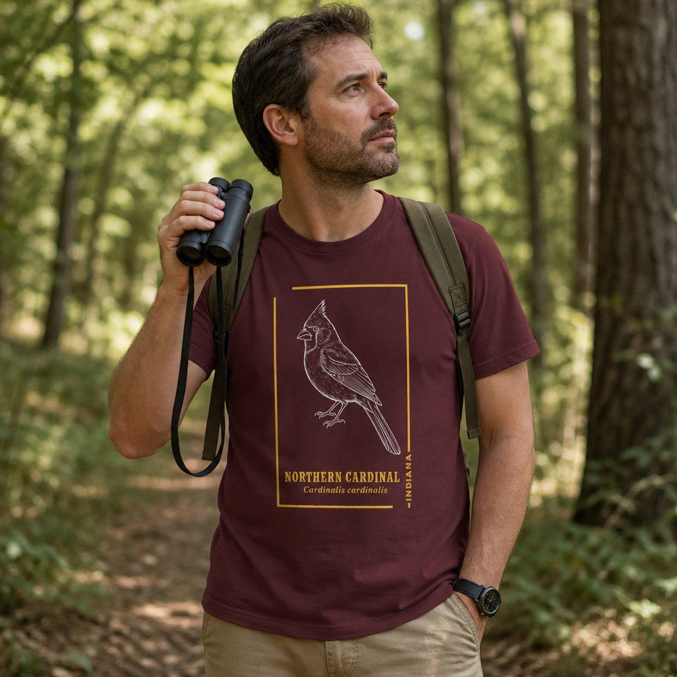 Man wearing a maroon Bella Canvas 3001 t-shirt featuring a vintage line art illustration of a Northern Cardinal with goldenrod frame and classic field guide typography, hiking in a lush green forest, ideal for birdwatchers and wildlife art lovers, by Crossroads Dandelion