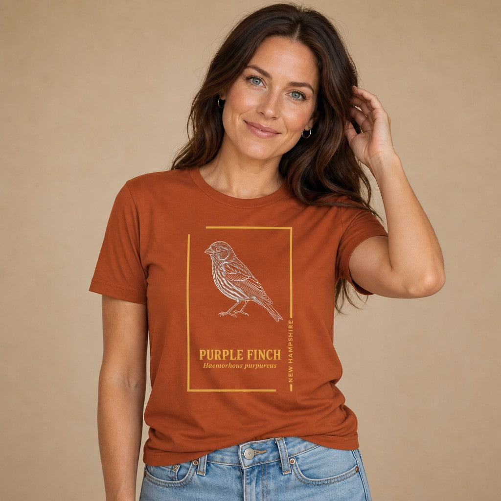 Woman wearing an autumn orange Bella Canvas 3001 t-shirt featuring a vintage-inspired hand-illustrated purple finch in detailed white ink with botanical field guide typography and gold frame, perfect for wildlife and New England nostalgia lovers, by Crossroads Dandelion