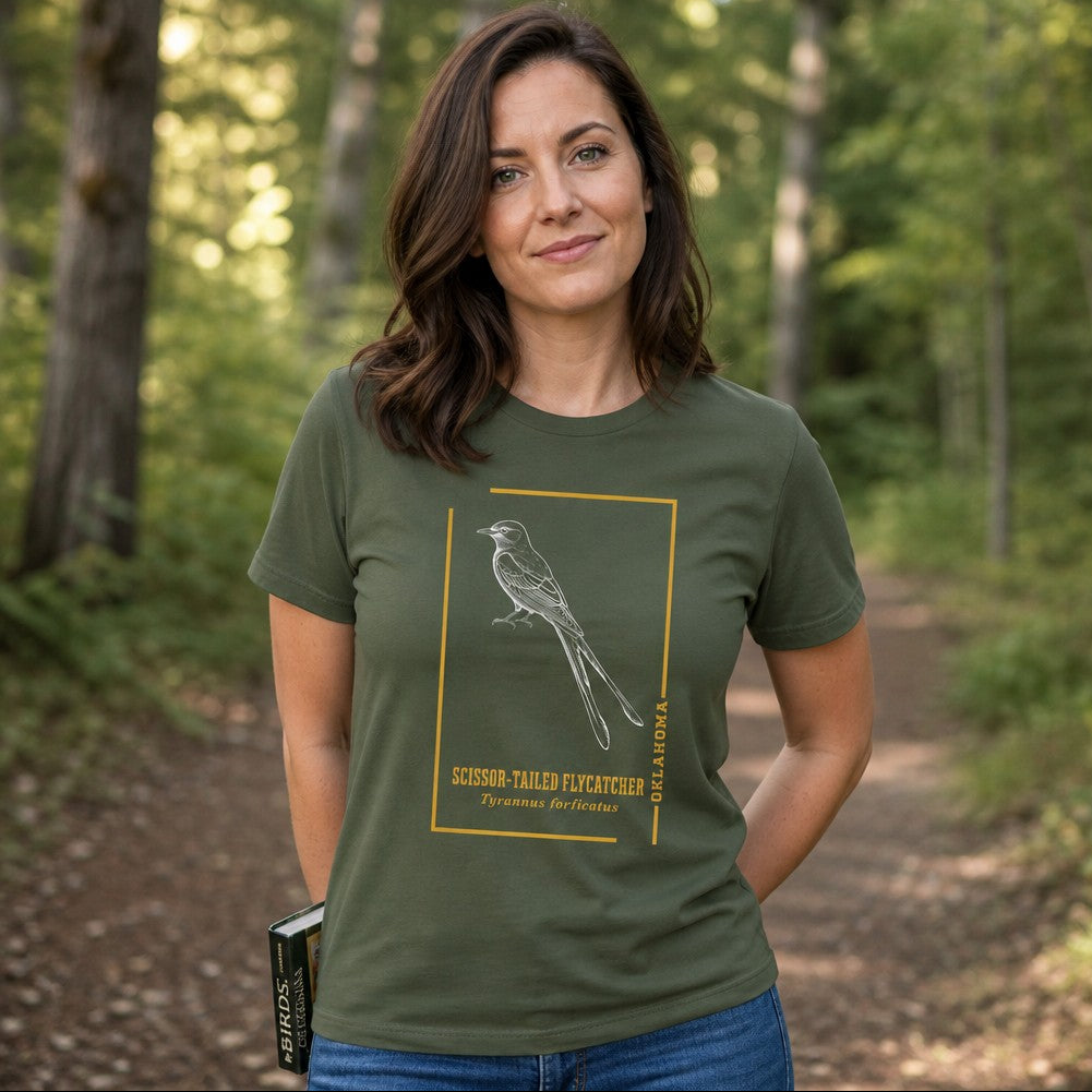 Woman wearing a military green Bella Canvas 3001 t-shirt featuring a vintage-inspired line art of a scissor-tailed flycatcher in a golden frame, perfect for bird lovers and nature enthusiasts, outdoor forest setting, by Crossroads Dandelion