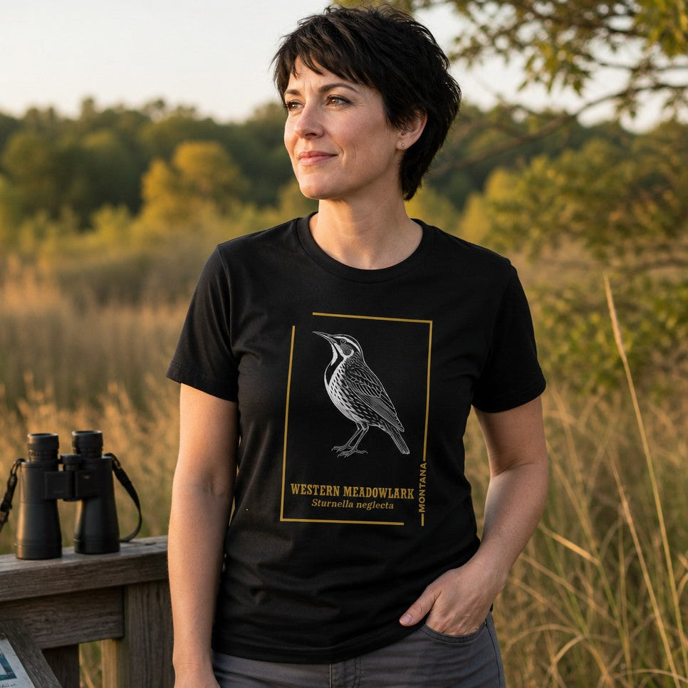 Woman wearing a black Bella Canvas 3001 t-shirt featuring a vintage line art etching of a Western Meadowlark framed in gold, classic field guide style, standing outdoors in a scenic nature setting, perfect for birdwatchers and nature art lovers, by Crossroads Dandelion
