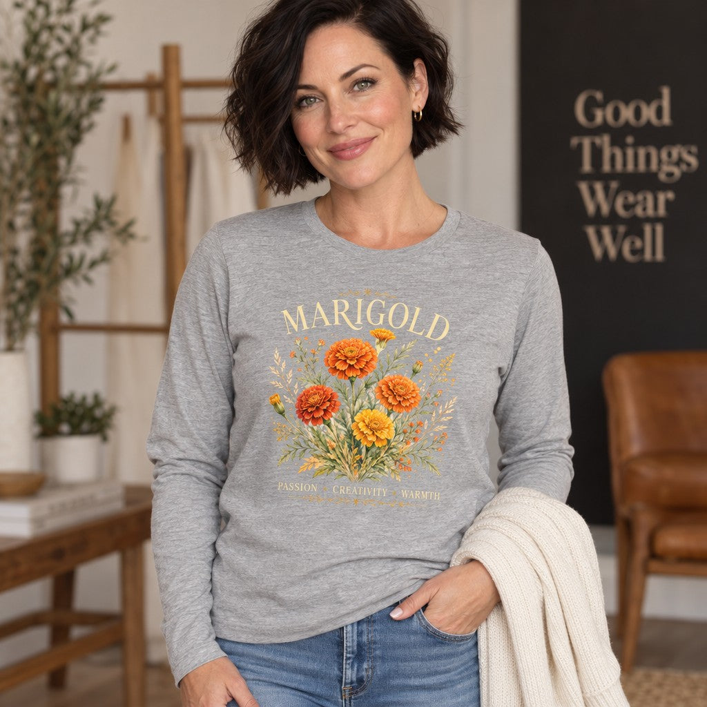 Woman wearing an athletic heather long sleeve Bella Canvas® 3501 shirt with a vintage watercolor marigold floral bouquet design, featuring arched serif MARIGOLD text and botanical symbolism for October birthdays, styled in a cozy home setting, for women and flower lovers, by Crossroads Dandelion