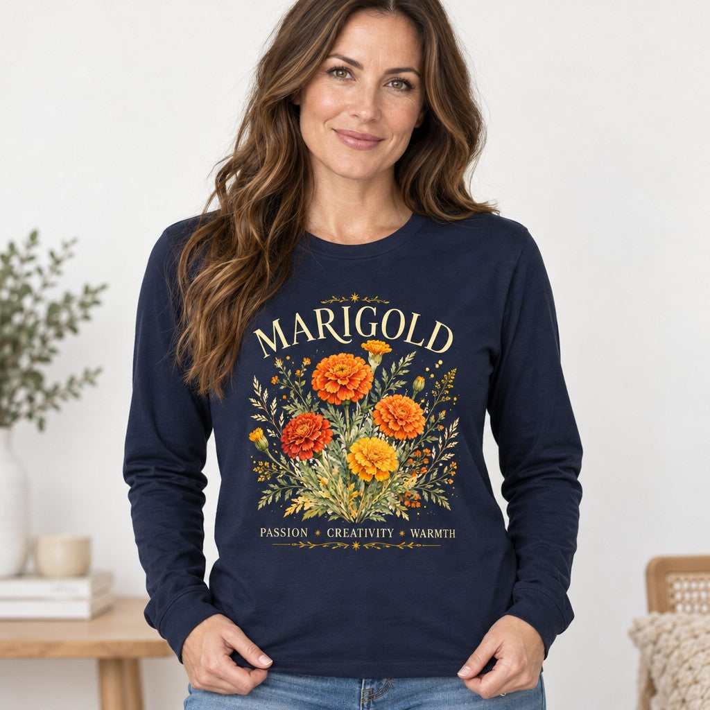 Woman wearing a navy Bella Canvas® 3501 long sleeve shirt with a vintage watercolor bouquet of gold and orange marigolds, botanical design with 'MARIGOLD' typography, ideal for October birthdays, gardeners, and flower-symbolism lovers, cozy indoor setting, by Crossroads Dandelion