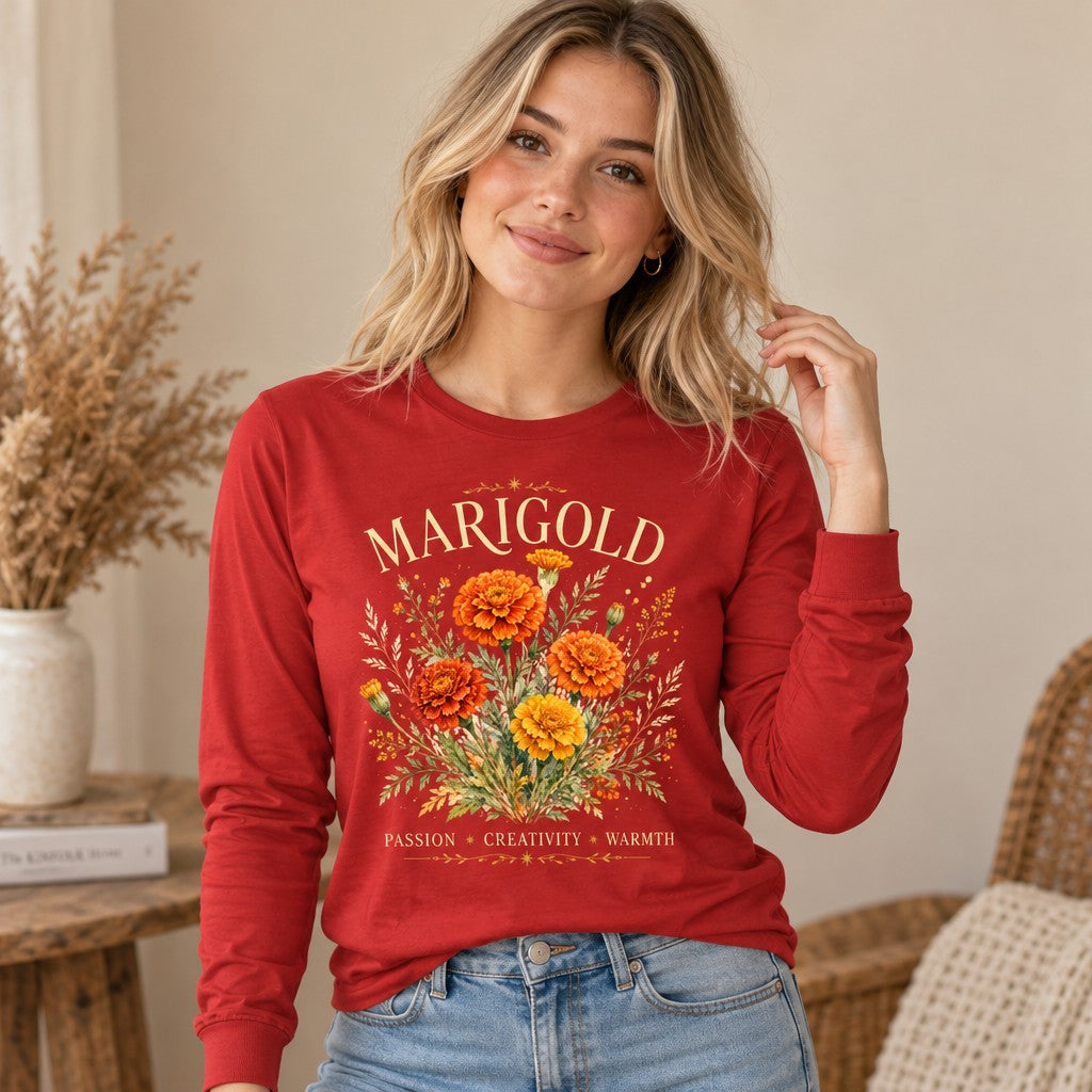 Woman in red Bella Canvas® 3501 long sleeve shirt with vintage watercolor marigold bouquet design, arched serif MARIGOLD text, PASSION CREATIVITY WARMTH message, cozy cottagecore indoor scene, perfect for October birthdays, flower symbolism fans, and gardeners, by Crossroads Dandelion