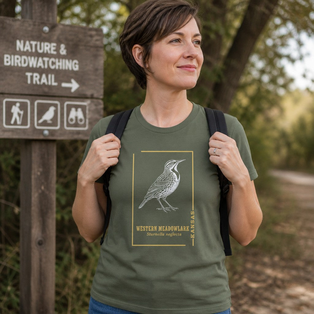 Woman wearing military green Bella Canvas 3001 t-shirt featuring vintage line art meadowlark illustration in golden frame with serif type and scientific name, standing at a nature and birdwatching trail, perfect for birdwatchers, prairie lovers, and outdoor enthusiasts, by Crossroads Dandelion