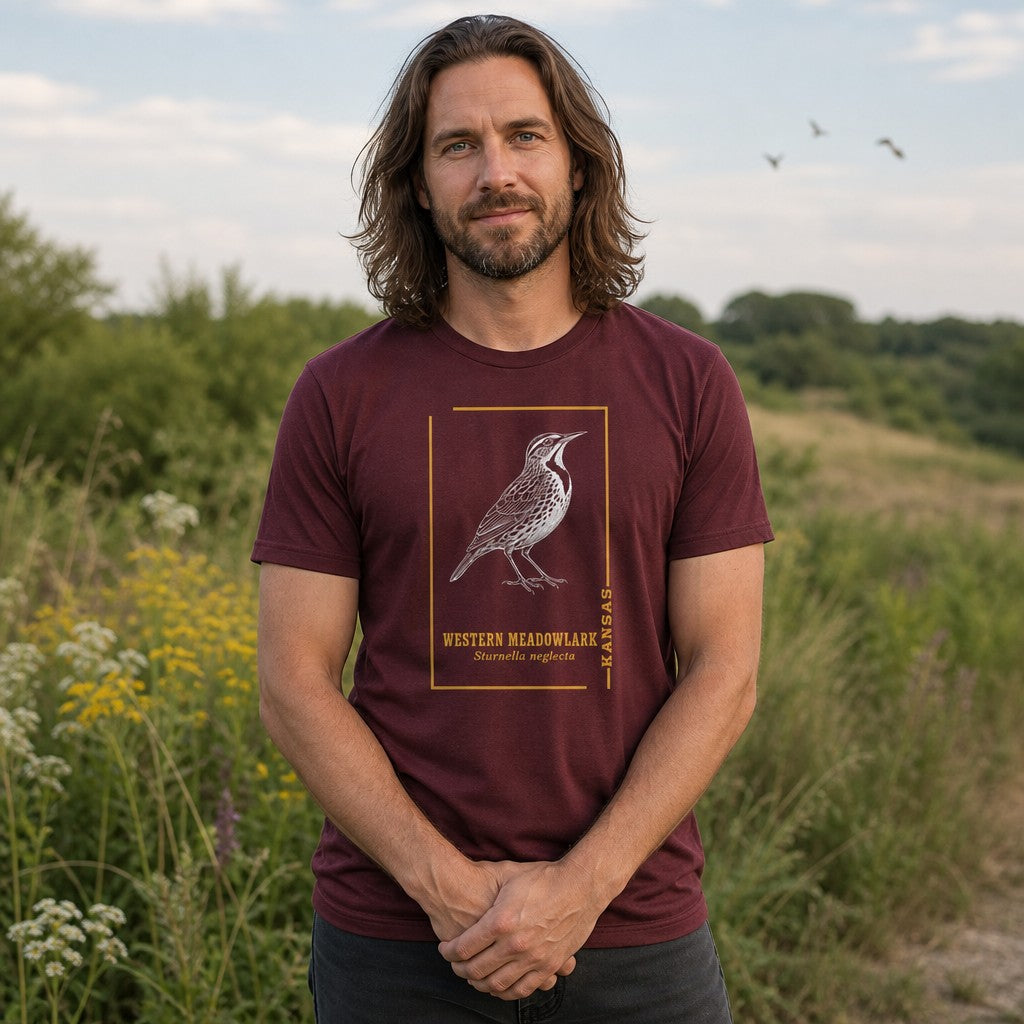 Man wearing a maroon Bella Canvas 3001 t-shirt featuring a black and white vintage meadowlark illustration framed in gold with classic serif typography, standing outdoors in a grassy field, perfect for bird watchers, nature and prairie nostalgia enthusiasts, by Crossroads Dandelion