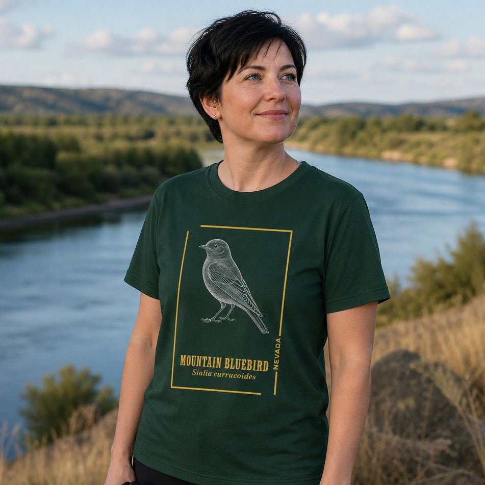 Woman wearing a forest green t-shirt with a vintage line art mountain bluebird design and scientific name, standing by a river with hills in the background, perfect for birdwatchers and nature lovers, by Crossroads Dandelion