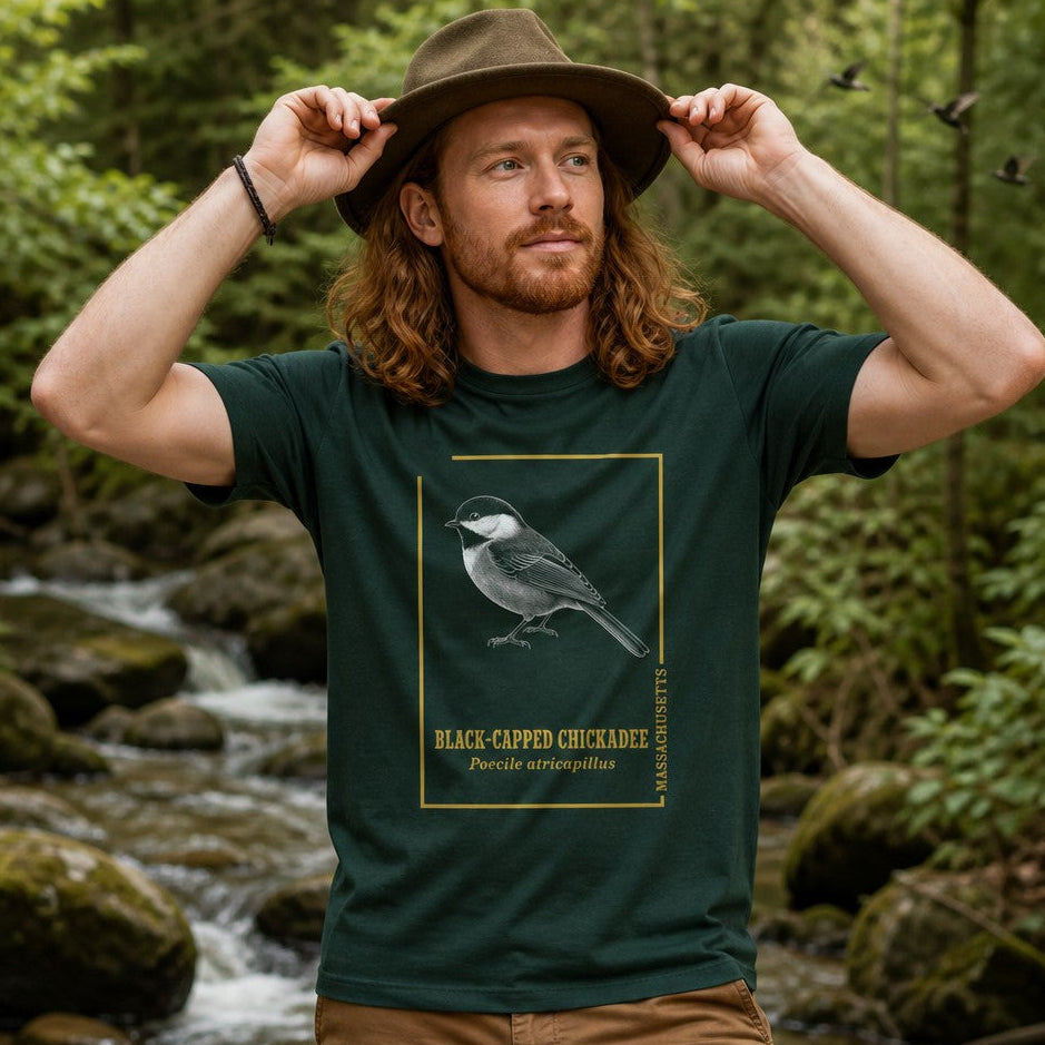 Man in forest green Bella Canvas 3001 t-shirt featuring a vintage black-capped chickadee naturalist engraving with golden frame and scientific text, hiking by a mountain stream in the woods, for nature and wildlife art lovers, by Crossroads Dandelion