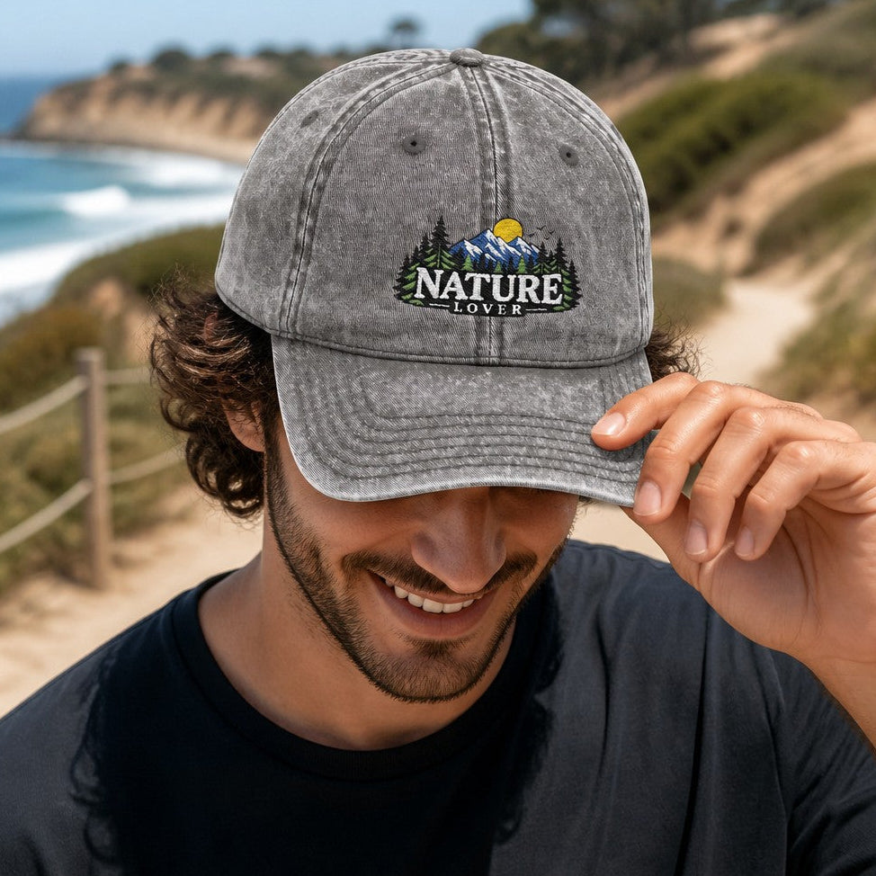 Man wearing a charcoal grey baseball cap with embroidered vintage nature lover design featuring mountains, trees, and sun, coastal trail background, perfect for outdoor enthusiasts and men, by Crossroads Dandelion