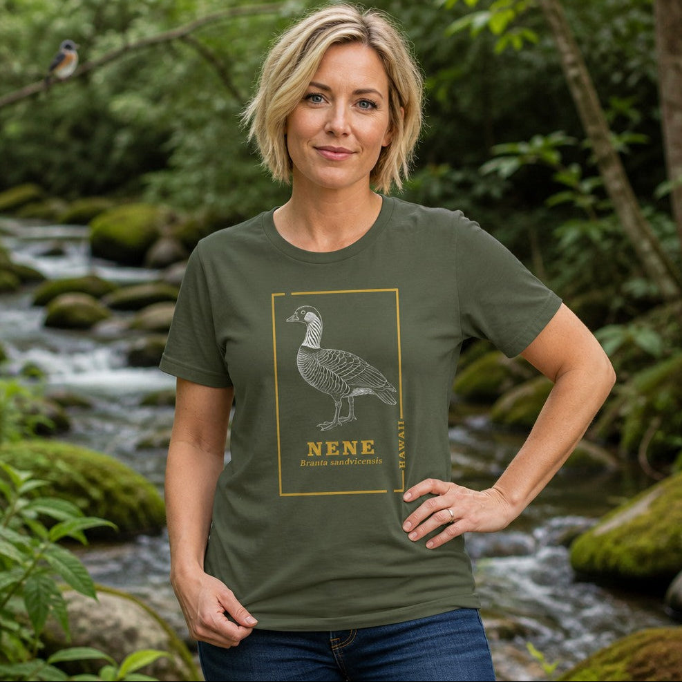 Woman wearing a military green Bella Canvas 3001 t-shirt with a vintage hand-drawn line art illustration of a nene goose and golden botanical frame, celebrating Hawaii's rare state bird, standing by a mossy stream in nature, ideal for birdwatchers and nature lovers, by Crossroads Dandelion