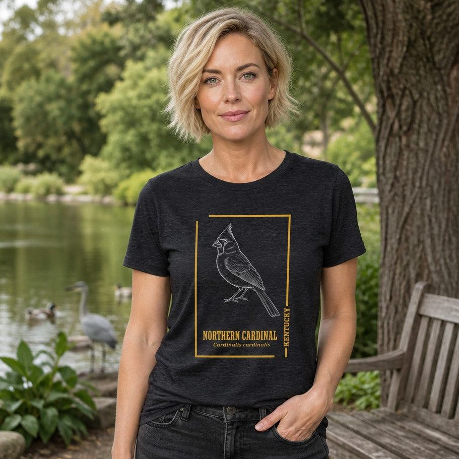 Woman wearing a dark grey heather Bella Canvas 3001 t-shirt featuring a vintage-inspired white line art illustration of a northern cardinal with gold accents and botanical typography, modeled outdoors by a pond for birdwatchers and nature lovers, by Crossroads Dandelion