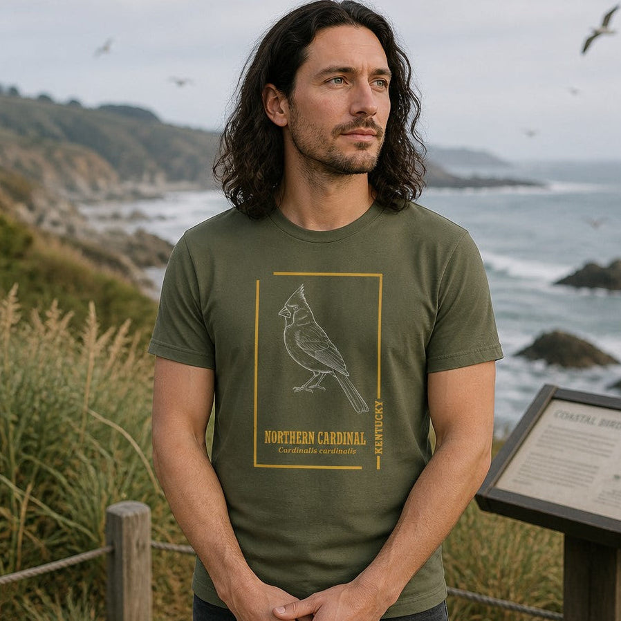 Man wearing a military green Bella Canvas 3001 t-shirt with a vintage-inspired hand-drawn white line illustration of a Northern Cardinal, bordered in gold with botanical typography, standing by a coastal trail overlook, perfect for birdwatchers and wildlife enthusiasts, by Crossroads Dandelion