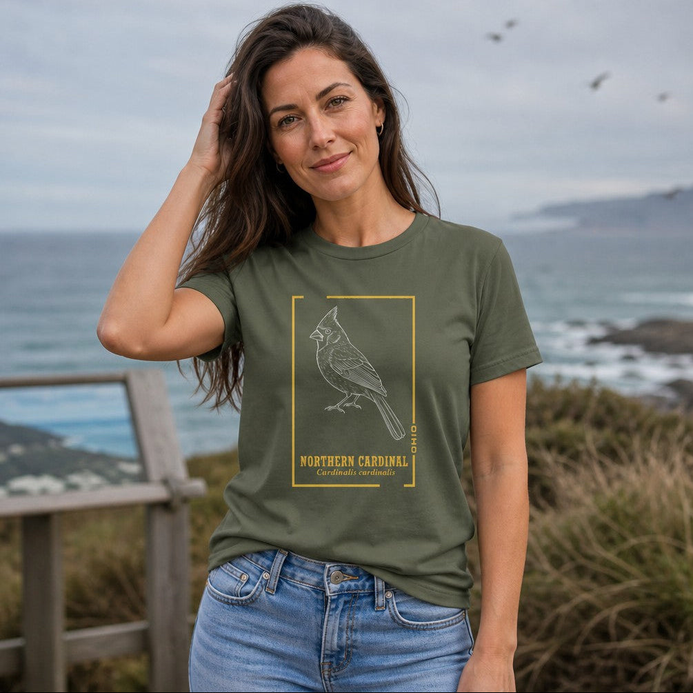 Woman wearing a military green Bella Canvas 3001 t-shirt featuring a vintage-inspired white line art illustration of a Northern Cardinal in a golden botanical frame with bold serif and italic Latin text, perfect for birdwatchers and vintage wildlife lovers, coastal outdoor setting, by Crossroads Dandelion