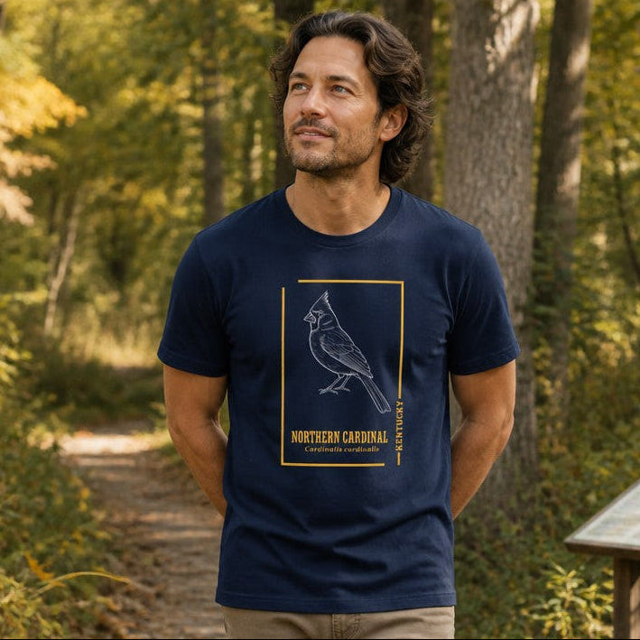 Man wearing navy Bella Canvas 3001 t-shirt featuring a vintage hand-drawn white line art illustration of a Northern Cardinal with gold botanical typography, minimalist field sketch style, standing on a woodland boardwalk, perfect for birdwatchers and nature lovers, by Crossroads Dandelion