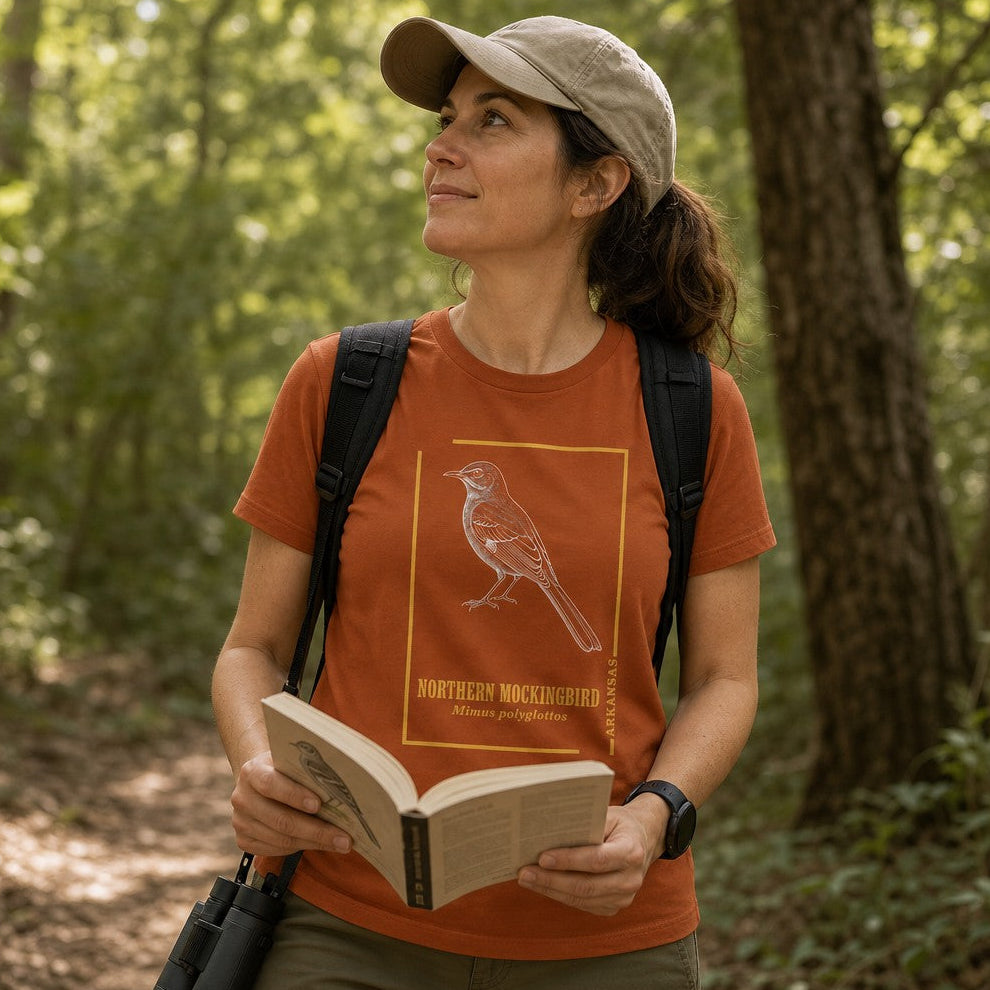Woman wearing an autumn Bella Canvas 3001 t-shirt featuring a vintage hand-rendered Northern Mockingbird illustration with a gold border, inspired by antique naturalist field guides, hiking in a sun-dappled woodland, perfect for birdwatchers and nature enthusiasts, by Crossroads Dandelion