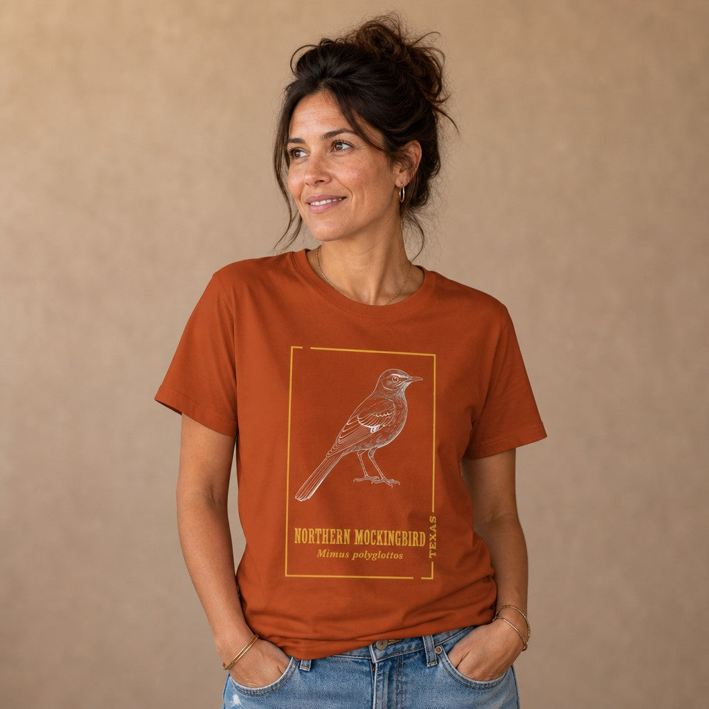 Woman wearing an autumn rust Bella Canvas 3001 t-shirt featuring a vintage-inspired white line art illustration of a northern mockingbird framed with gold accents and classic serif text, minimalist indoors setting, perfect for birdwatchers and Texas natives, by Crossroads Dandelion