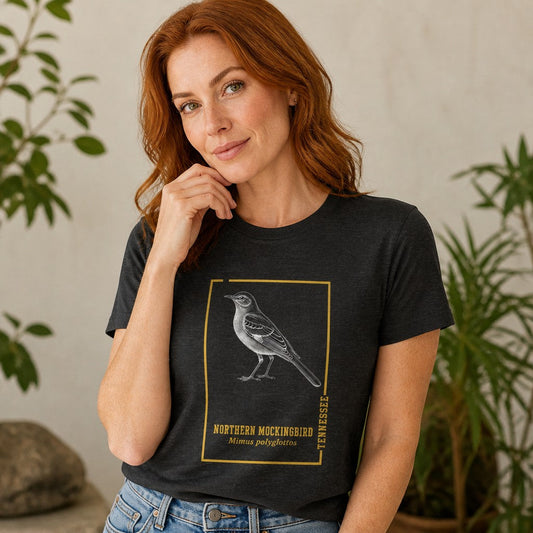 Woman wearing a dark grey heather Bella Canvas 3001 t-shirt with a vintage northern mockingbird engraving inside a golden botanical frame, antique field guide style, ideal for birdwatchers and Appalachian wildlife enthusiasts, indoor setting, by Crossroads Dandelion