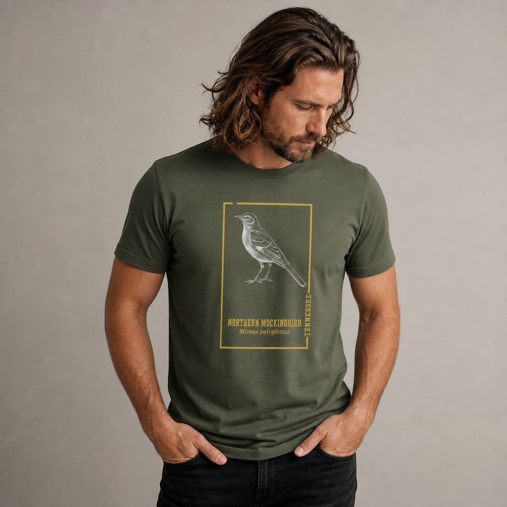 Man wearing military green Bella Canvas 3001 t-shirt featuring a vintage-style engraving of a northern mockingbird inside a golden botanical frame, field guide inspired bird illustration ideal for birdwatchers and Appalachian wildlife lovers, by Crossroads Dandelion