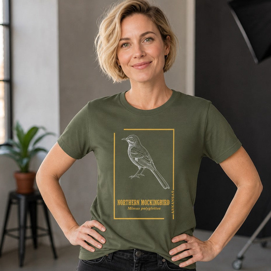 Woman wearing a military green Bella Canvas 3001 t-shirt featuring a vintage hand-drawn Northern Mockingbird within a golden frame, inspired by antique field guides, perfect for birdwatchers and nature lovers, studio setting, by Crossroads Dandelion