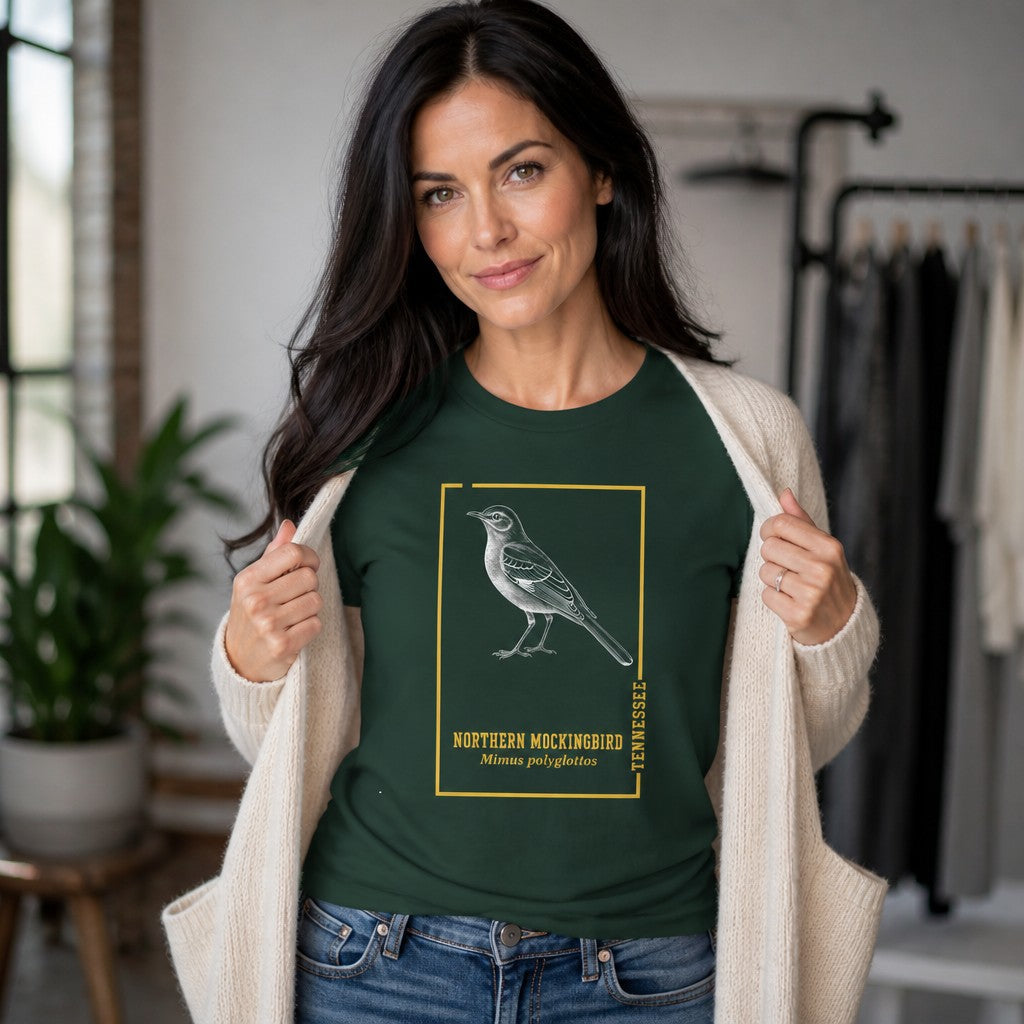 Woman wearing a forest green Bella Canvas 3001 t-shirt featuring a vintage-style engraved northern mockingbird in a golden botanical frame, antique field guide aesthetic, perfect for birdwatchers and Appalachian wildlife lovers, photographed indoors, by Crossroads Dandelion