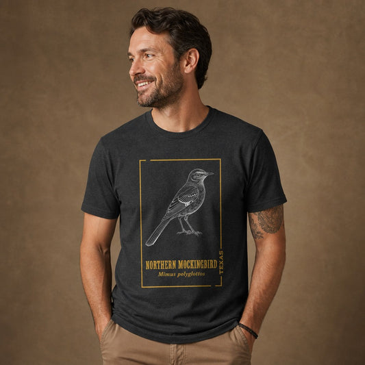Man wearing dark grey heather Bella Canvas 3001 t-shirt featuring a vintage hand-drawn white line art illustration of a northern mockingbird, framed in gold accents with classic serif text, perfect for Texas natives, birdwatchers, and lovers of nostalgic field guide art, by Crossroads Dandelion